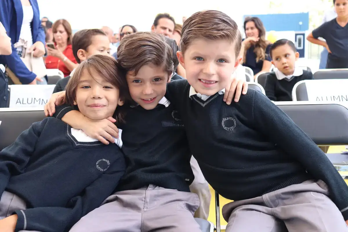 Tres estudiantes sonrientes en uniforme escolar sentados juntos durante un evento en el colegio.