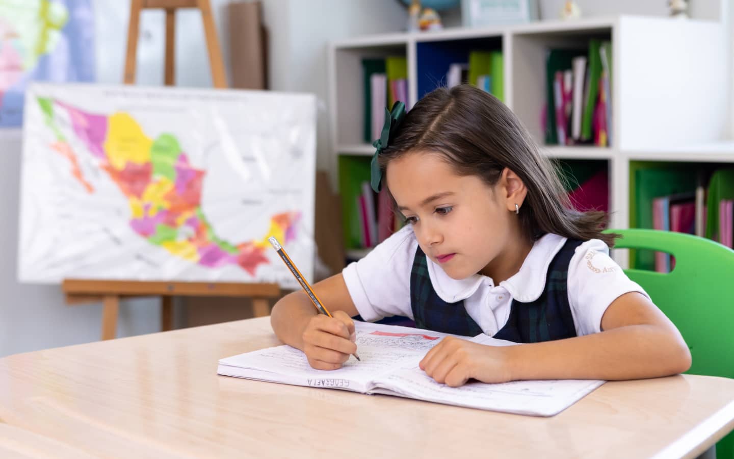 Niña en uniforme escolar escribiendo en un cuaderno en un aula con un mapa de México y estantes con libros al fondo.