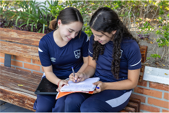 Dos estudiantes femeninas en uniforme azul sentadas en un banco de madera revisando y escribiendo en unos cuadernos al aire libre.