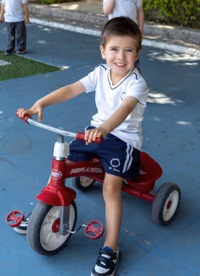 Niño sonriente sentado en un triciclo rojo sobre un pavimento azul en un parque.