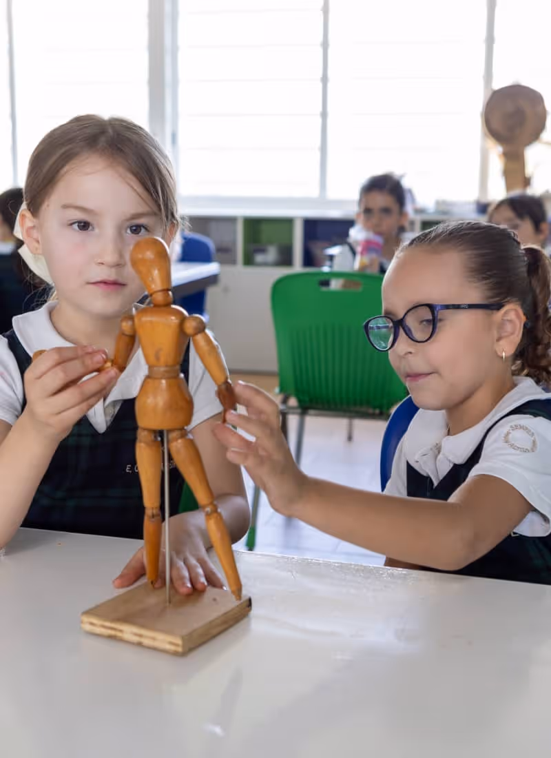 Dos niñas en uniforme escolar interactuando con una figura de madera articulada en un salón de clases.