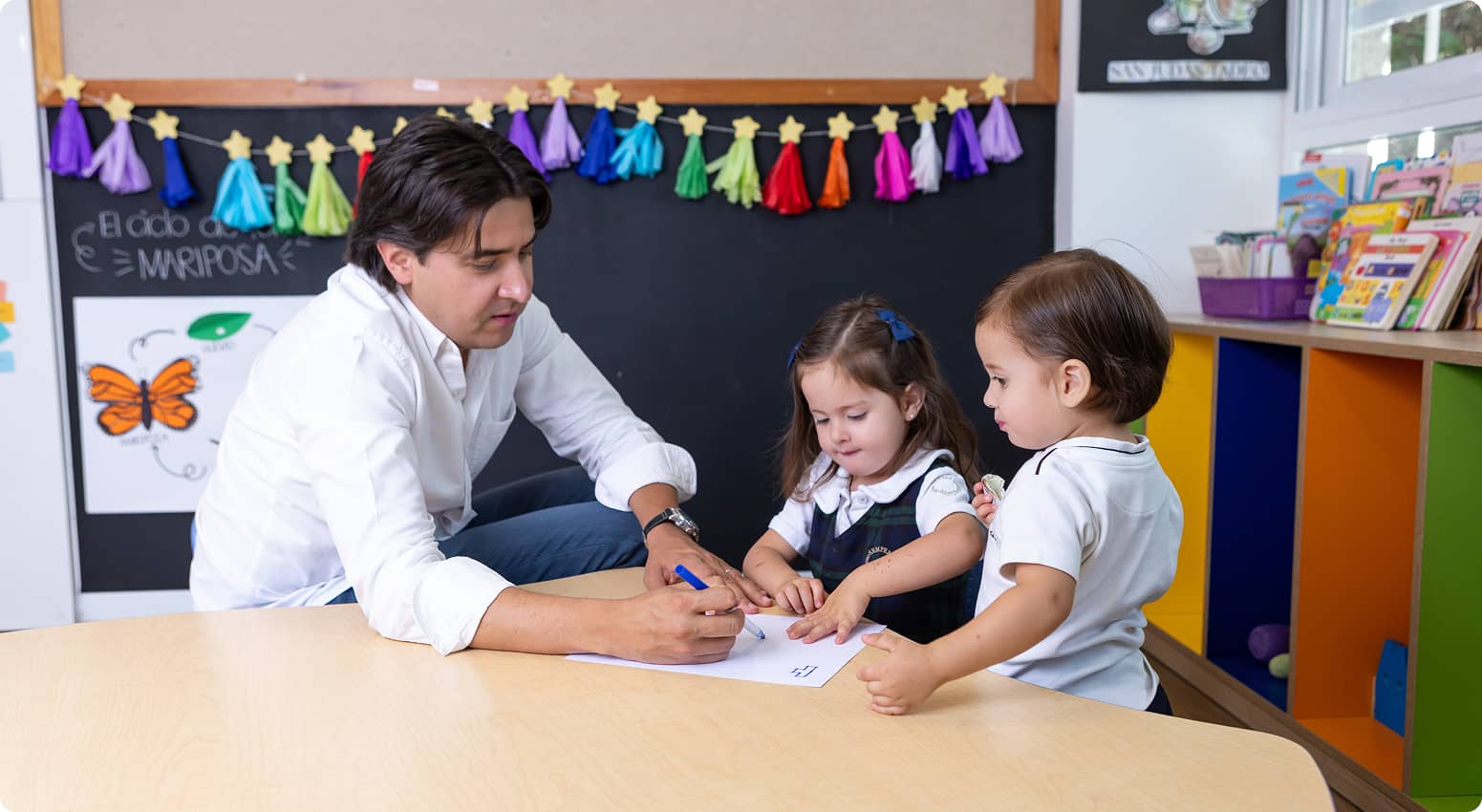 Adulto enseñando a dos niños pequeños a escribir en una hoja en un aula decorada con guirnaldas de colores y dibujos de mariposas.