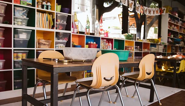 Mesas con sillas amarillas en un aula con estantes organizados con materiales escolares.
