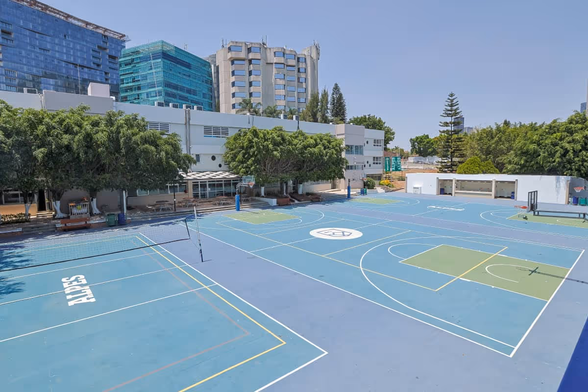 Un complejo deportivo al aire libre con canchas de baloncesto y voleibol, además de zonas para otros deportes, rodeado de árboles y adyacente a edificios modernos: un entorno ideal para programas educativos y fomentar el liderazgo.