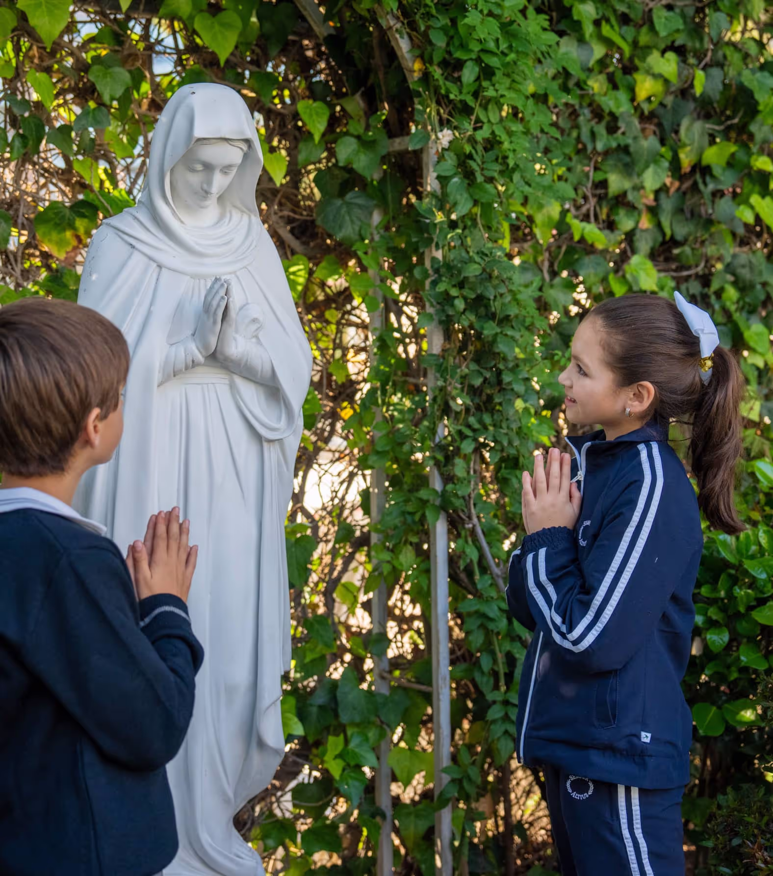 Niño y niña vestidos con uniforme escolar rezando frente a una estatua blanca de la Virgen María rodeada de vegetación.